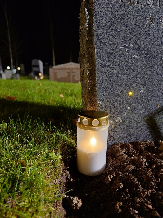 Gravljus vid en gravsten på en kyrkogård under natten. Gräs och jord syns i förgrunden.