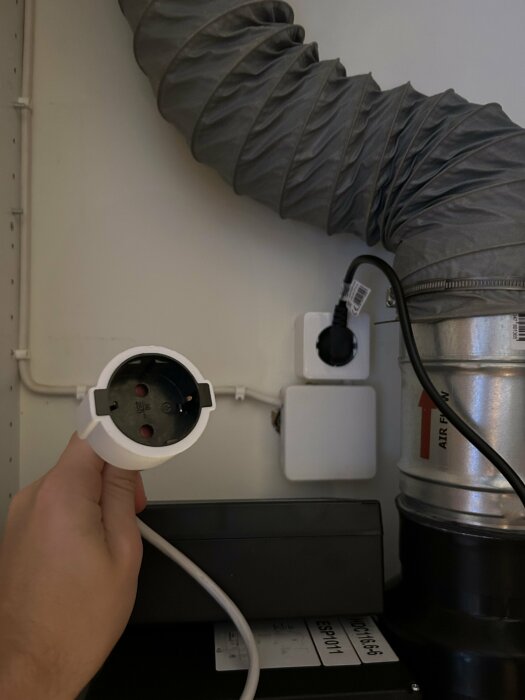 Hand håller i en vit kontakt framför en grå ventilationsslang och metallrör, bredvid ett eluttag med en svart kabel inkopplad.