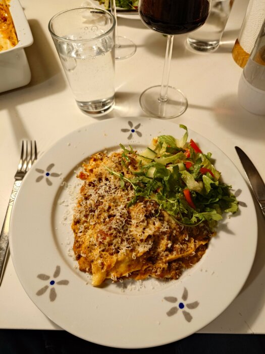 Lasagne med parmesan och svartpeppar, serverad med en grönsallad på en tallrik, glas rött vin och vatten bredvid.