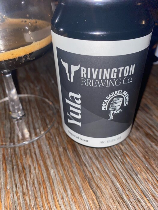 Burk med Yula Imperial Stout från Rivington Brewing Co. och ett glas delvis fyllt med öl på en träyta.