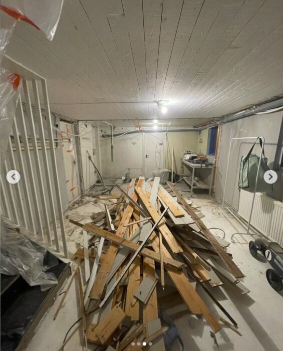 Hög med rivningsmaterial inklusive träpaneler på golv i källare under renovering, med arbetsutrustning i bakgrunden.