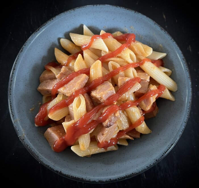 Pasta penne med korvgryta och vitkål, toppad med ketchup på en grå tallrik.