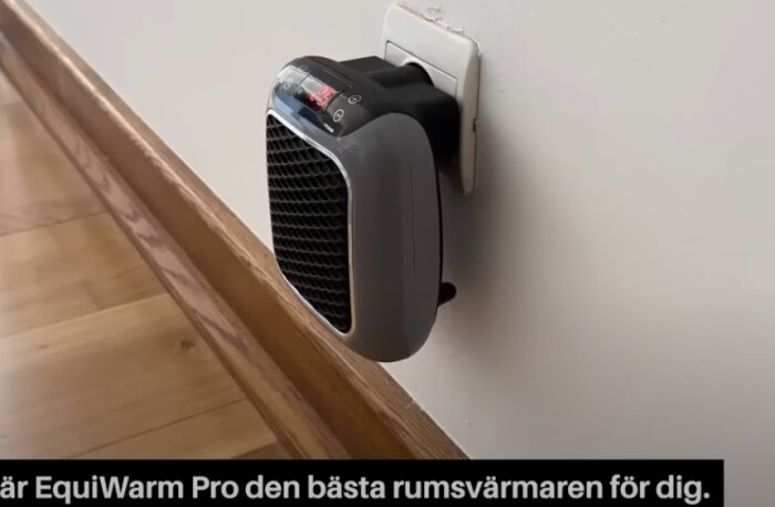 En EquiWarm Pro rumsvarmare är inkopplad i ett eluttag på en vit vägg, med ett trägolv nedanför.