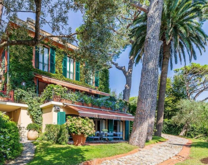 Villa i Italien omgiven av grönska och stora träd, med stenväg i förgrunden och blommor på terrassen.