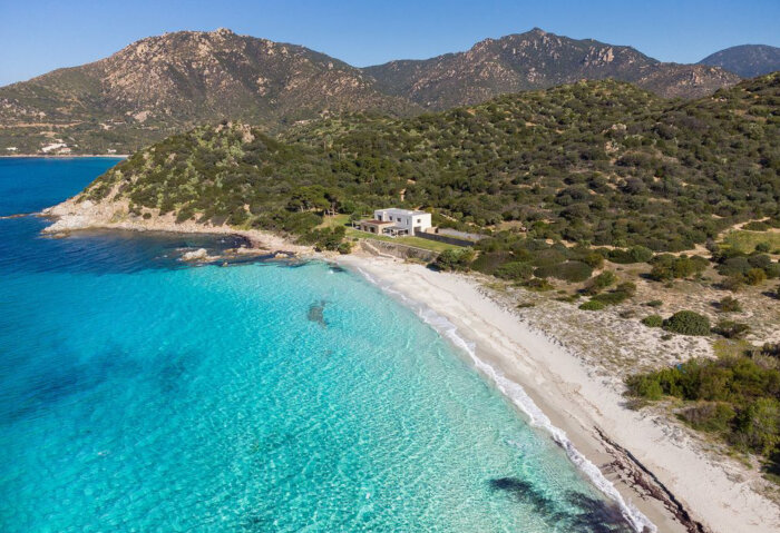 Vy över en villa vid en vit sandstrand och turkost hav i Villasimius, Sardinien, omgiven av gröna berg och skog.