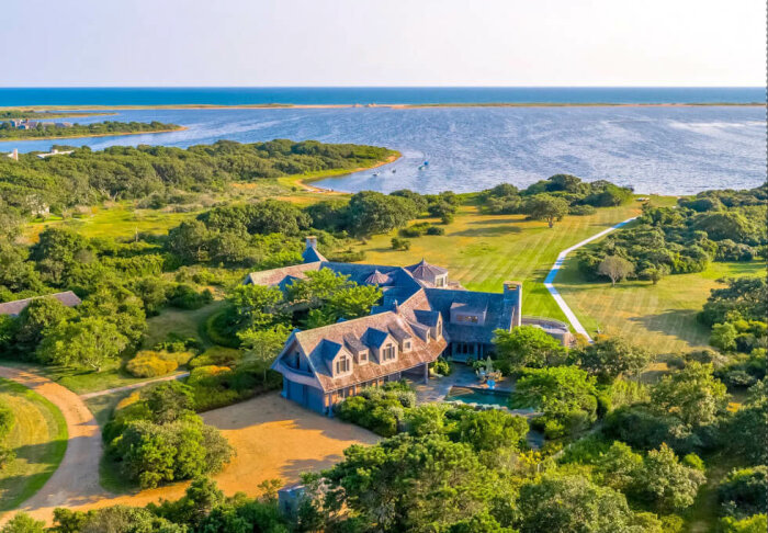 Flygbild av ett stort hus och omgivande grönska vid havet, med en bred gräsmatta och skog.