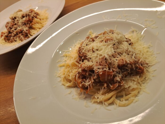 Två tallrikar spaghetti med köttfärssås, toppade med parmesanost, ligger på ett träbord.