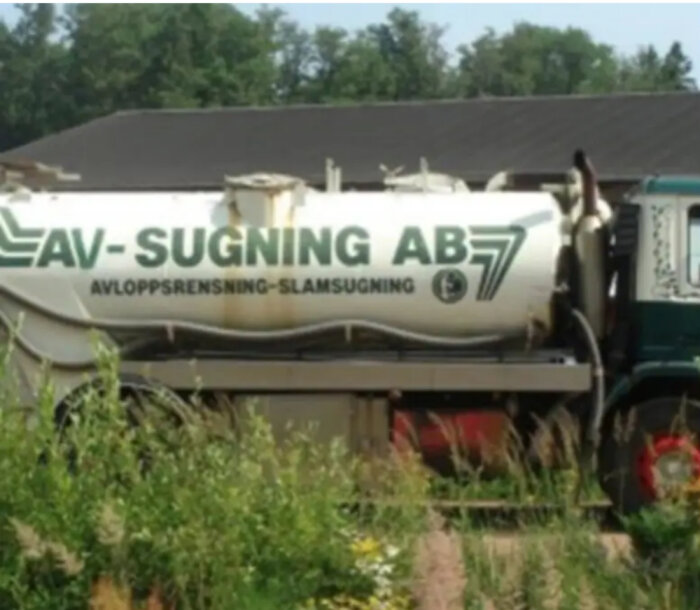 Tankbil med texten "AV-SUGNING AB", avloppsrensning och slamsugning, parkerad i grönt område framför en byggnad.