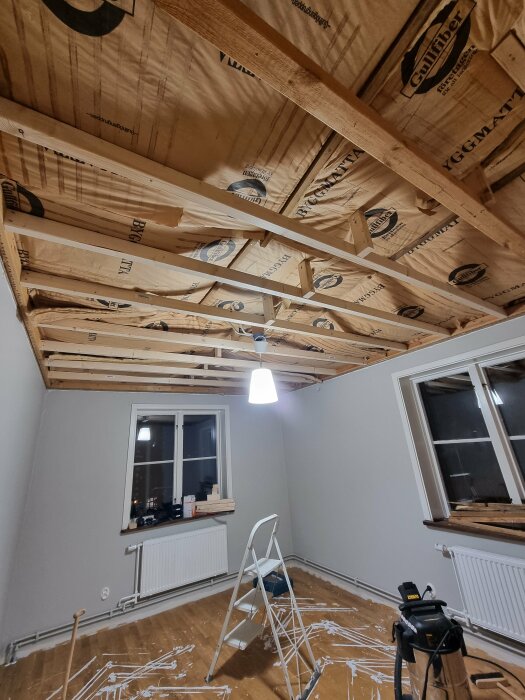 Reglar och isolering installerade i taket på ett renoveringsprojekt. Stege och verktyg syns i ett rum med trägolv och fönster.