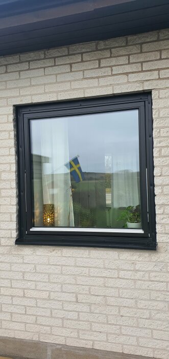 Fönster med svart ram i tegelvägg, reflekterar en svensk flagga och inredning med lampa och krukväxt inuti.