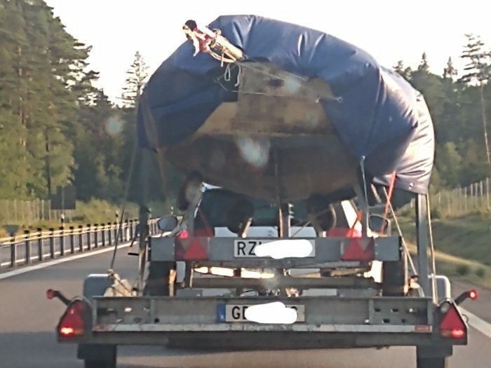 Biltrailer transporterar täckt båt på landsväg, flera fordon synliga i bakgrunden.