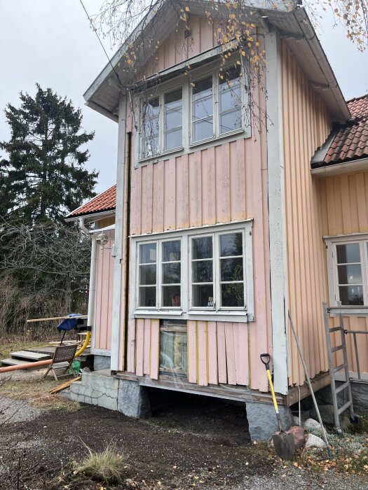 Utbyggnad med rosa fasad och synlig grund, verktyg lutade mot väggen, träd i bakgrunden. Planerad renovering pga dålig lukt och fuktproblem.