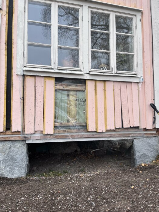 Utbyggnad med rosa träpanel och synlig trossbotten, antyder problem med dålig lukt och fuktig isolering. Fönster ovanför och synlig grund.