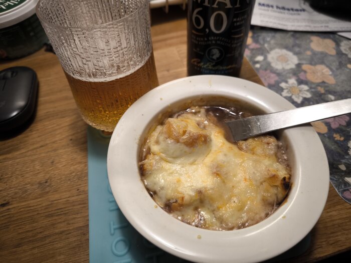 Fransk löksoppa med gratinerad ost och baguette, serverad i en skål med sked, och ett glas öl bredvid på ett träbord.