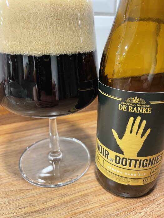 Glas och flaska av ölen De Ranke Noir de Dottignies på ett träbord. Mörk öl med skum i glaset och etikett på flaskan.