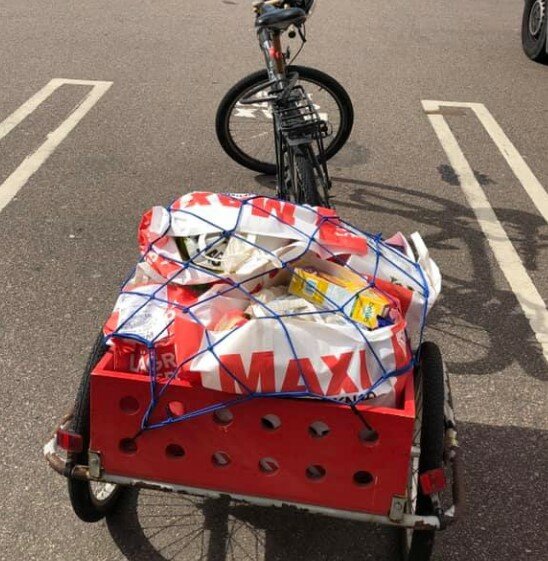 Cykel med en släpvagn fylld med varor i plastkassar, varav många är från en butik med namnet Maxi, fastspända med blått nät.