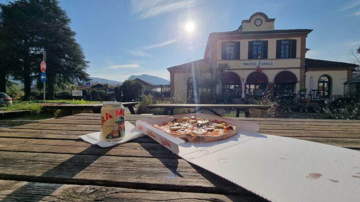 Pizza och öl på ett träbord utanför en byggnad med italiensk flagga under solig himmel.