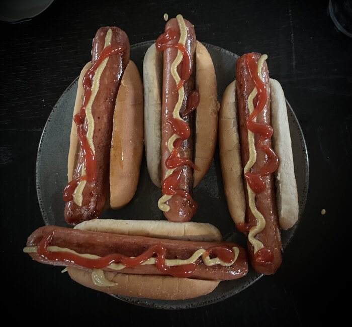 Fem grillkorvar i bröd med ketchup och senap på tallrik.