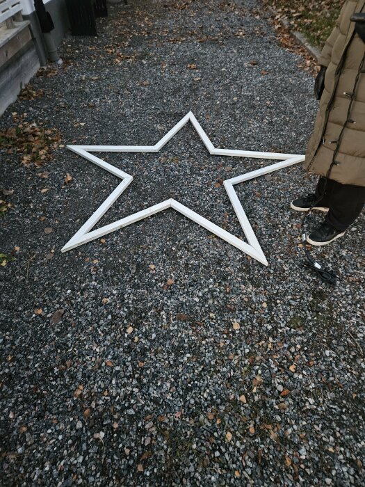 Vit träram i form av en stjärna ligger på grusgång, omgiven av höstens löv och en person står bredvid i vinterkläder.