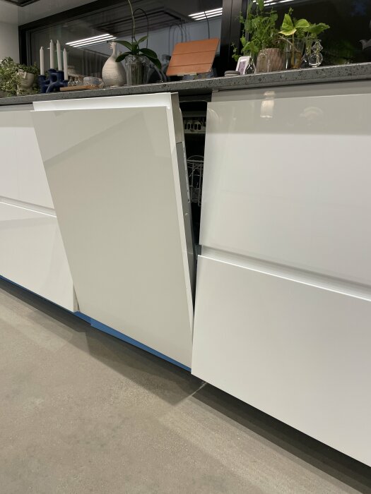 Vitt IKEA-kök med delvis öppen kökslucka visar en Miele diskmaskin bakom. Svart bänkskiva med krukväxter och föremål.