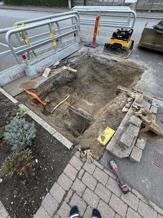 Grävd grop i trottoar med avstängningshinder och verktyg, förberedelse för byte av vattenrör, med handske och verktygslåda i förgrunden.