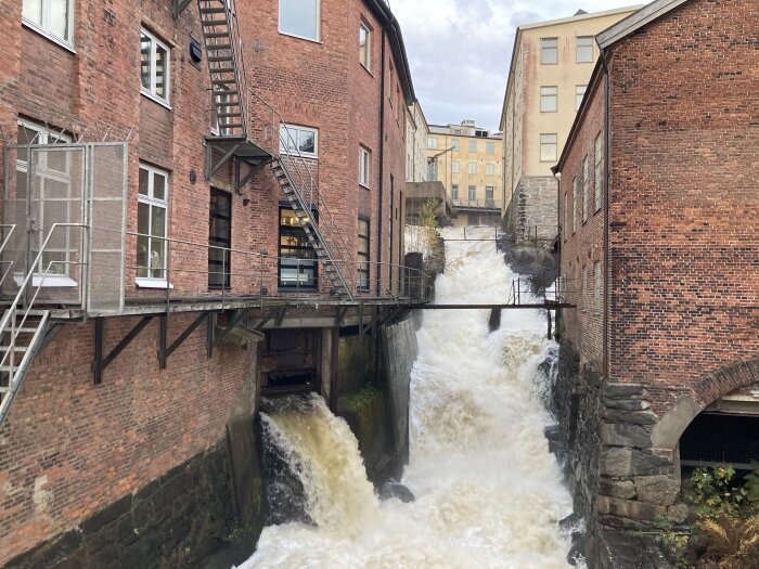 Vatten forsar ut från husgrunden mellan tegelbyggnader, liknande en mindre fors, vilket kan indikera fuktproblem i äldre fastighet.