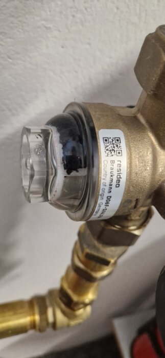 Närbild på en tryckregulator för inkommande vatten. Synligt är filter och kopplingar. Märken och etiketter kan ses på regulatorn.