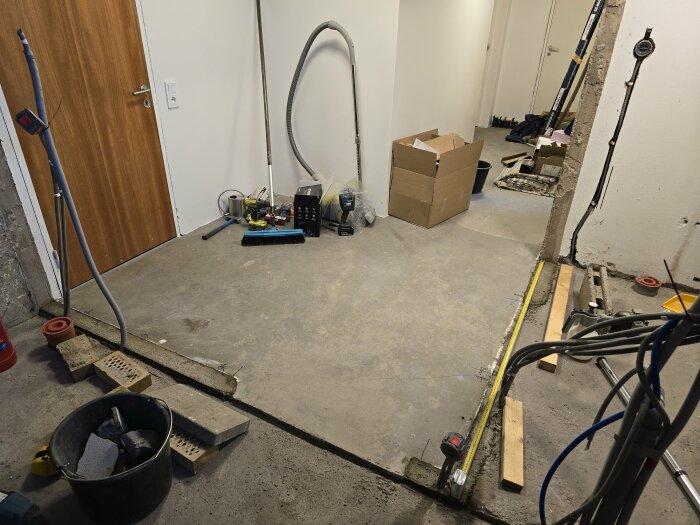 Renovering av tvättstuga; betonggolv, hål i väggen, verktyg, slangar och material utspridda. Kartong och uppstädningsredskap synliga.