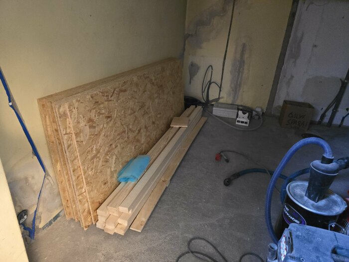 OSB-skivor och träreglar staplade mot en vägg i ett rum under renovering, med dammsugare och byggmaterial på golvet.