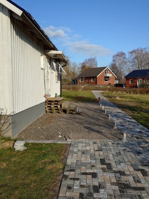 Gångväg av marksten bredvid ett hus med staplade träpallar och tegelstenar, omgiven av en grön gräsmatta och blå himmel.