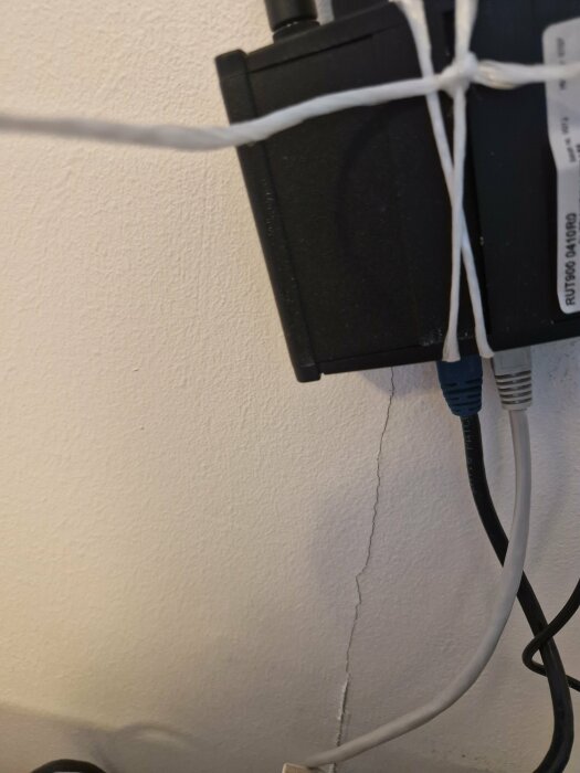 Router monterad på vägg med nätverkskablar och förstärkare, knuten med vitt snöre. Spricka synlig på väggen bakom.