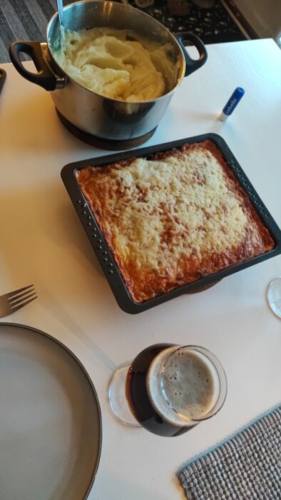 Gryta med potatismos, ugnsform med korvgratäng och ett glas mörk julöl på ett bord.