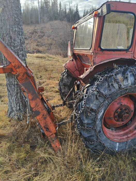 Traktor med snökedjor på däcken och vajer monterad på sidan, vid ett träd på gräsbeklätt område.