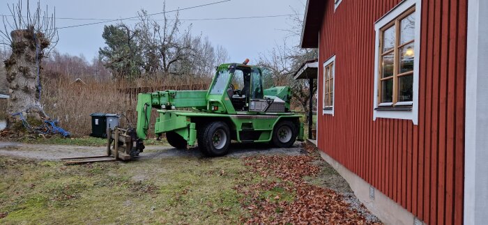 Grön teleskoplastare parkerad bredvid en röd byggnad med trädgård och träd i bakgrunden.