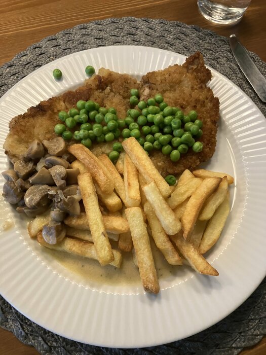 Schnitzel med gröna ärtor, pommes frites och svampsås på en vit tallrik.