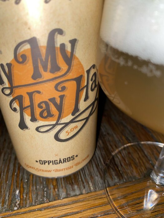 En burk Oppigårds My My Hay Hay öl med 5% alkoholhalt bredvid ett glas öl på ett träbord.