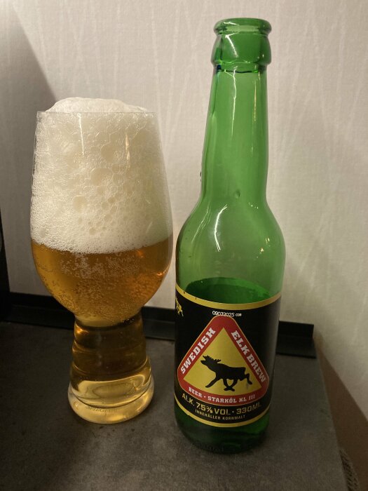 Grön flaska och glas öl med skumkrona, etikett visar "Swedish Elk Brew". Alkolhalt 7,5%, flaskstorlek 330 ml.