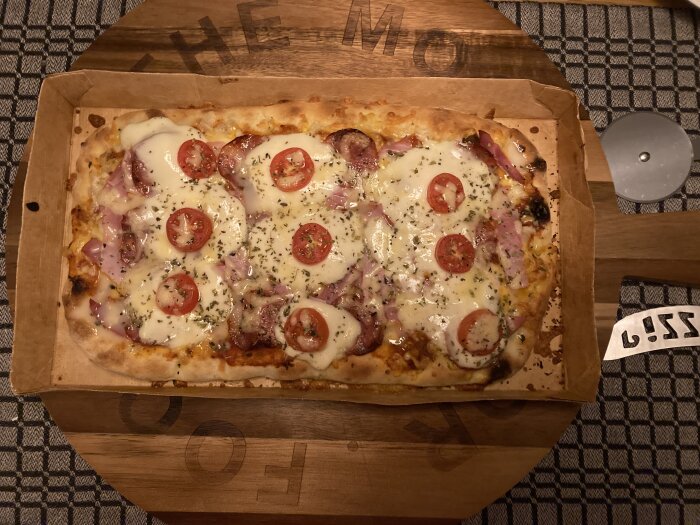 Rektangulär pizza med salami, mozzarella, skinka, tomater och pizzaost på ett runt serveringsbräde.