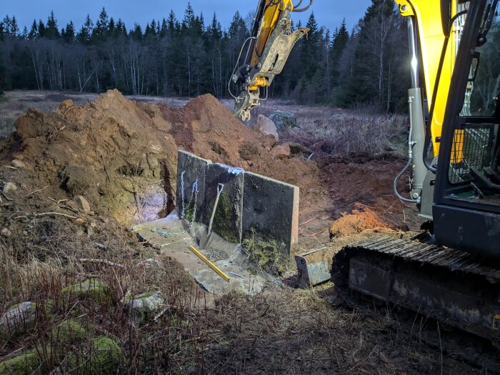 En grävmaskin vid en byggarbetsplats där L-stöd placeras i marken, omgiven av jordhögar och skog i bakgrunden.