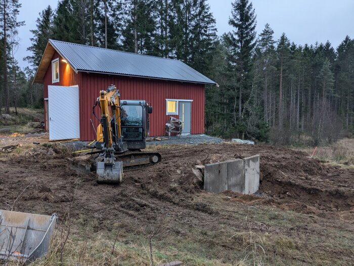 Grävmaskin vid rödmålat garage, l-stöd placerade i marken, omgiven av träd och natur.