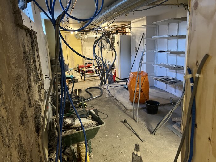 Renovering av källare med blå nätverksslangar, hyllor och verktyg synliga. Flytspacklad yta och arbetshörn. Ventilationsrör i taket.