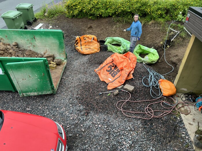 Man sorterar skräp på en gårdsplan med en grön container och sopsäckar i olika färger, en person står bland kablar och verktyg.