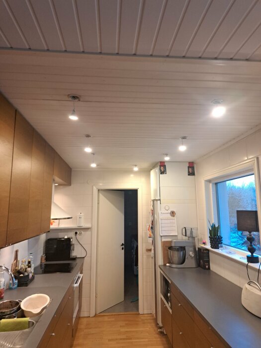 Kök med vit paneltak, LED-spotlights installerade. Träskåp och grå bänkskiva synliga. Flera spotlights i taket, vissa flimrar.