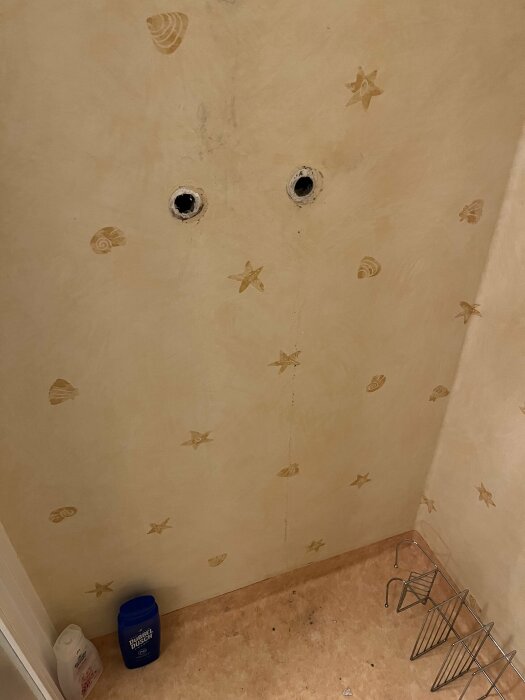 Vägg med stjärn- och snäckmönster där två hål för en borttagen blandare syns, samt tecken på fukt under en matta.
