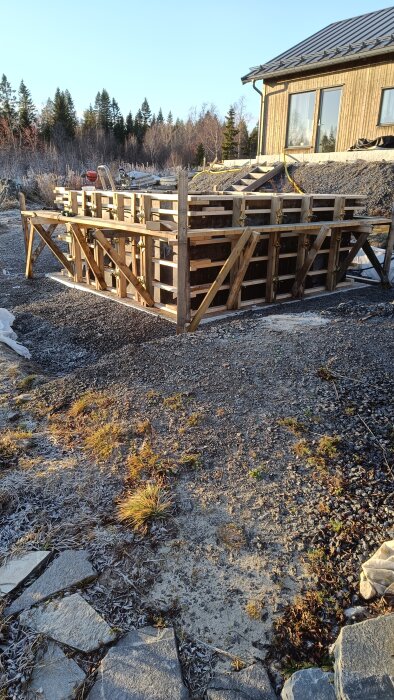 Byggarbetsplats med träform för gjutning, omgiven av grus, vid ett hus; redo för betonggjutning efter formbyggnad och armering.