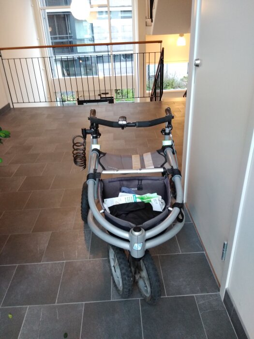 Terränggående rollator med tre hjul, varav framhjulet består av två förskjutna hjul, står i en modern hall med trist kakelgolv.