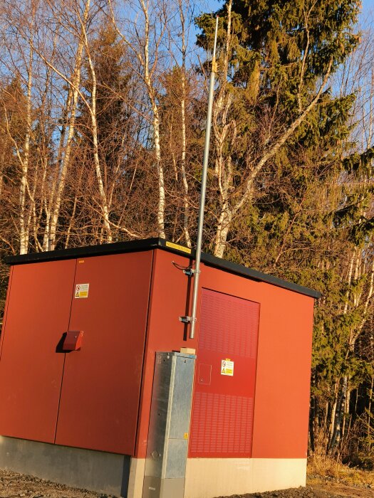 Röd transformatorstation med antenn och varningsskyltar, omgiven av träd, i ett bostadsområde.