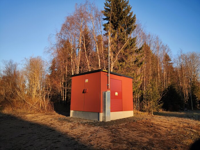 Röd transformatorstation med antenn i soligt skogslandskap.