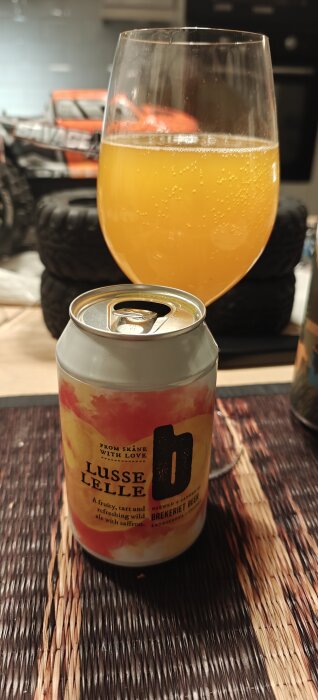 Burk och glas med orangefärgad öl, etiketten "Lusse Lelle" från Brekeriet Beer synlig. I bakgrunden syns leksaksbilens hjul.