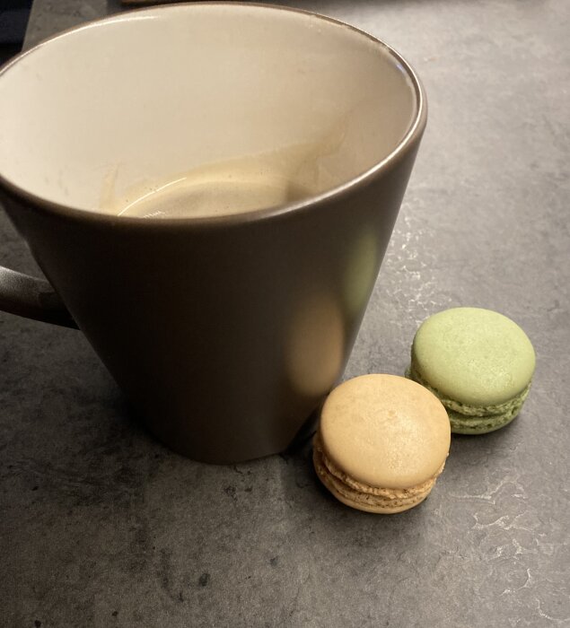 En mugg med kaffe bredvid två macarons, en grön och en beige, placerade på en mörk bänkskiva.